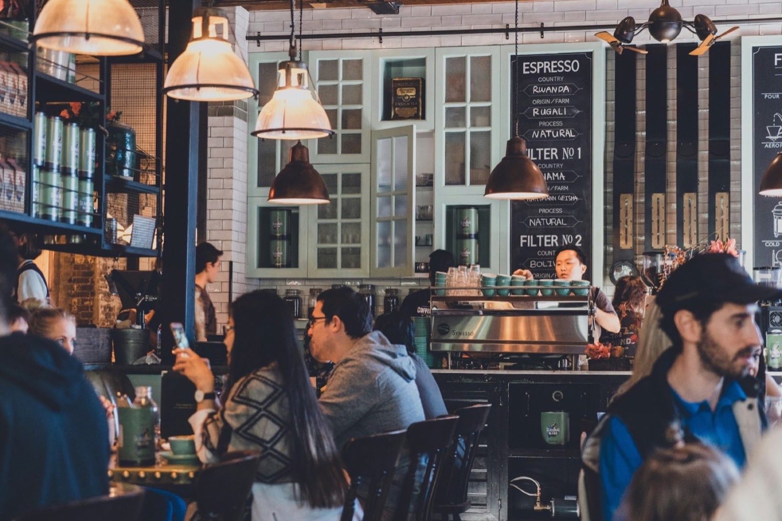 A busy coffee shop interior with an espresso bar, tiled wall and people at tables