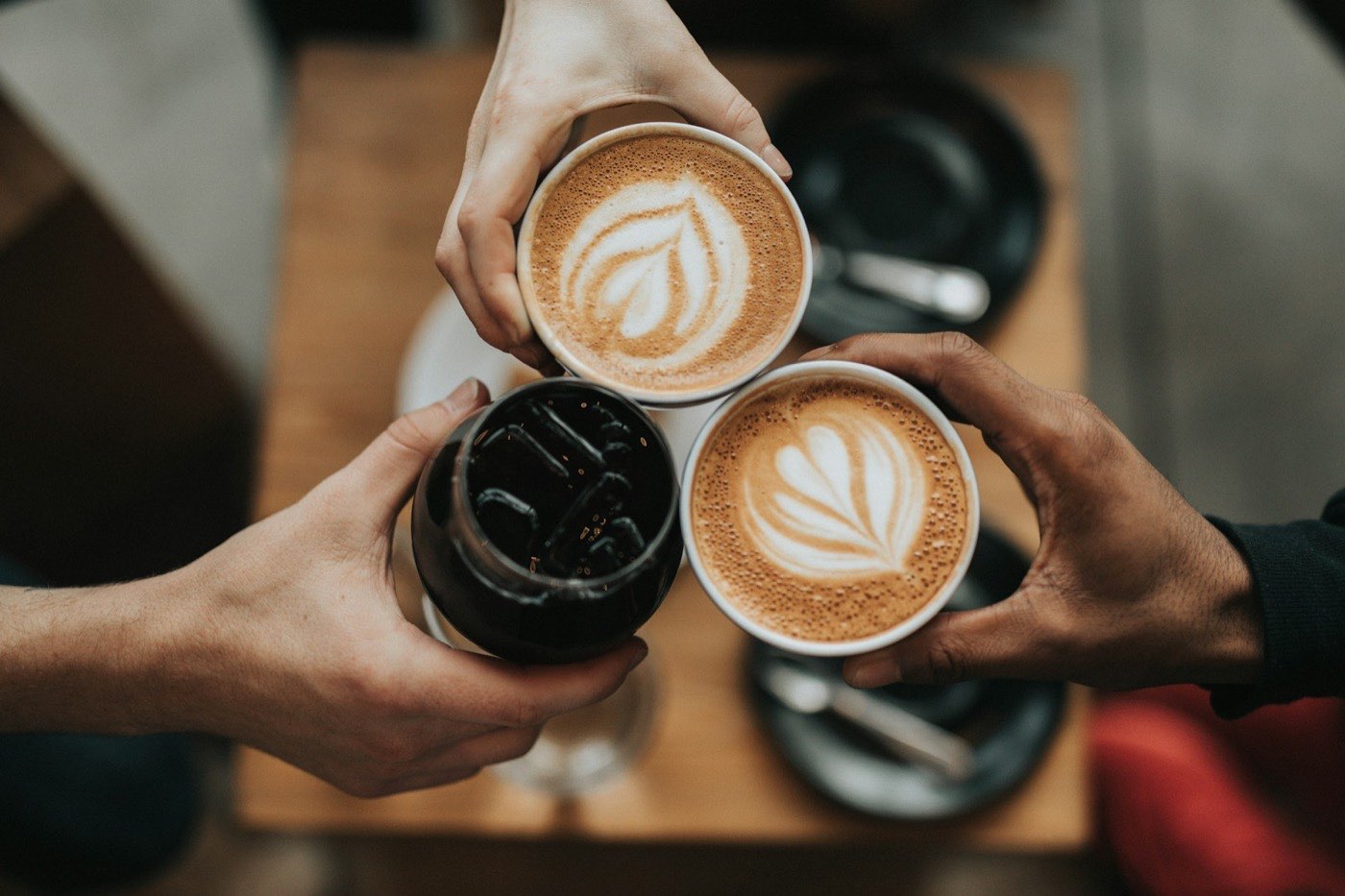 Three hands holding lattes with leaf latte art and an iced coffee over a wooden table