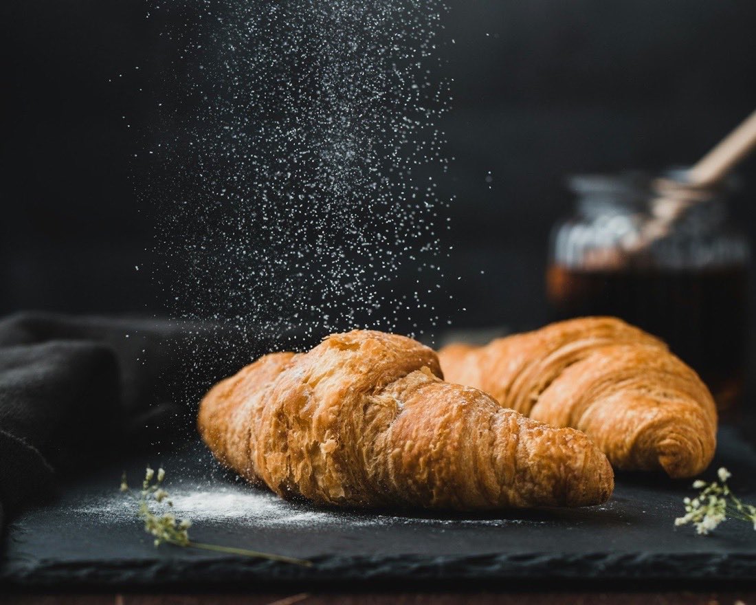 Two croissants on a slate board with icing sugar falling over them